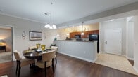 The Benjamin's two-bedroom, two-bathroom residence in Chamblee, GA presents a thoughtfully designed single-floor layout that balances open communal spaces with private retreats. The heart of the home is an open-concept living, dining, and kitchen area anchored by rich hardwood-style flooring. The kitchen features warm cherry cabinetry, a double-basin sink, a breakfast bar peninsula with pendant lighting, and black appliances, including a full-size refrigerator. It opens seamlessly to the dining area, which is illuminated by an elegant chandelier, and flows into the generous living room. The living room is finished with crown molding, a ceiling fan, large windows, and French doors that invite abundant natural light and provide access to an outdoor space with tree-lined views. Both bedrooms are carpeted and well-sized, offering ample room and natural light through large windows. The primary bedroom includes an en-suite bath with a wide vanity, white cabinetry, tile flooring, and a walk-in shower area. The second bathroom is similarly appointed. Tile flooring carries through the kitchen and bathrooms, while the neutral color palette throughout the residence creates a cohesive and welcoming ambiance.