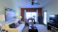 This single-story space centers around a well-appointed living room that combines comfort and style through a thoughtful mix of design elements. A striking terracotta-red accent wall serves as the focal point, framing a large window dressed with white curtains that allows generous natural light to fill the room. A second window on the adjacent wall further brightens the space, enhancing its open, airy feel.

The ceiling features a classic multi-blade ceiling fan with an integrated light fixture, providing both air circulation and ambient lighting. Neutral carpet flooring runs throughout, contributing to a warm and inviting atmosphere. The living area is furnished with a plush white sofa accented with decorative throw pillows in bold patterns, complemented by a patterned armchair in rich tones. A dark leather ottoman serves as a central coffee table, topped with a decorative tray.

Finishing touches include a large indoor plant in the corner, a table lamp adding warm ambient light, and wall art that adds a splash of color. Together, these elements create a cohesive, livable space with a balance of bold accents and neutral tones.