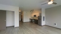 This contemporary single-floor space offers a clean, open-concept design that seamlessly integrates the living and kitchen areas into one cohesive layout. The kitchen is outfitted with a full suite of stainless steel appliances, including a refrigerator, range, microwave, and dishwasher, complemented by modern dark cabinetry and light countertops. Track lighting above the kitchen provides focused illumination, enhancing both function and ambiance. The flooring throughout is a light-toned hardwood-style material that lends warmth and continuity across the space. Crisp white walls and smooth ceilings contribute to a bright, airy atmosphere that feels open and uncluttered. A hallway visible from the main living area leads to additional rooms, suggesting a practical and well-organized floor plan. A utility panel is neatly integrated into the wall, maintaining the space's clean aesthetic. The overall design strikes a balance between modern minimalism and everyday functionality, making it a versatile and inviting environment suited for a variety of lifestyles.