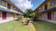 This single-story unit is part of a two-story courtyard-style building characterized by its bold yellow exterior, brick base, and red-accented doors arranged symmetrically around a lush central green space. The courtyard features mature trees, tropical palms, and planted garden beds centered around a decorative stone feature, creating a tranquil and well-maintained outdoor environment.

Inside, the unit showcases a refined interior defined by polished concrete floors that lend an industrial yet sophisticated character to the space. The dining area serves as a focal point, anchored by a striking round green marble table paired with woven rattan chairs atop a natural fiber rug. A large-scale framed artwork with subtle, neutral tones adorns the white walls, complementing the understated palette throughout.

A sleek sliding barn door with matte black hardware provides a stylish transition between rooms, adding a contemporary architectural detail. Recessed lighting and well-placed HVAC vents maintain a clean ceiling plane, while textured walls contribute warmth and depth. An adjacent room, visible through a set of vintage-style doors, displays additional wall art and continues the curated aesthetic. The overall ambiance is modern and artfully composed, balancing industrial elements with refined, eclectic furnishings.
