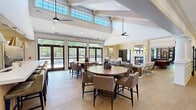 Located at 1900 Barclay Blvd in Princeton, NJ, this generously sized single-floor clubhouse offers an impressive array of amenity spaces designed for both social gatherings and quiet retreats. The centerpiece is a soaring great room with vaulted ceilings accented by exposed wood beams and clerestory windows that flood the space with natural light. Multiple round dining tables with upholstered chairs provide ample seating, complemented by ceiling fans and large glass doors that frame views of the surrounding outdoor deck and greenery. A stunning double-sided marble fireplace anchors the lounge area, flanked by gold wall sconces and elegant millwork, creating a refined yet welcoming atmosphere. The kitchen features custom light wood cabinetry with a marble-look backsplash, sleek black hardware, and modern appliances — ideal for catering and entertaining. A sophisticated game room offers wainscoted walls in rich gray tones, a full billiards table, an upright piano, and built-in shelving. A cozy dining nook with sage-green cabinetry and garden views provides an intimate setting. Wide-plank hardwood flooring flows throughout, unifying each space. Classic six-panel white doors and crown molding complete the polished, upscale aesthetic of this exceptional clubhouse.