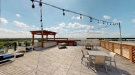 This well-appointed building offers a thoughtfully designed set of amenities centered around comfort, convenience, and outdoor living. The rooftop deck is a standout feature, finished with wide-plank decking and furnished with multiple dining and lounge seating arrangements. String lights are strung overhead, creating an inviting atmosphere for both daytime and evening use. A warm wood-toned pergola anchors one corner of the deck, complemented by a built-in grilling station nearby. The deck offers expansive panoramic views of the surrounding landscape, including a nearby body of water and open greenery beneath open skies.

Interior common areas are finished with light wood-look tile flooring and clean white walls, lending a bright, contemporary feel throughout. A well-lit corridor features curated abstract artwork, adding a refined aesthetic touch, and leads to a glass-panel entry door connecting to the outdoor spaces. The fitness center is accessible via full-length glass doors, offering visibility into a well-equipped workout space that includes treadmills, weight machines, free weights, and stability equipment — all within a mirrored, naturally lit interior. A dedicated package room houses an Amazon Hub locker system, providing a secure and convenient solution for parcel retrieval.