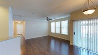 This single-floor space offers a clean, functional layout with a warm and inviting ambiance throughout. The main living area is a standout feature, showcasing rich, dark hardwood-style flooring that contrasts beautifully with the bright white walls and warm yellow accent tones. A ceiling fan is centrally positioned, adding both practicality and a subtle decorative touch to the room. Natural light pours in through a set of large windows fitted with blinds, creating a bright and airy atmosphere while offering easy light control. A sliding glass door adds an additional source of natural light and provides convenient access to the outdoors. Crown molding detail along the ceiling adds a refined architectural element to the living space. The open feel of the main room is enhanced by clean sight lines and smooth transitions into adjoining areas, visible through a hallway that connects to the rest of the unit. The overall design is straightforward and well-maintained, emphasizing comfort, functionality, and a neutral palette that offers versatility for a variety of interior styles.