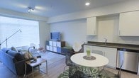 This single-floor space offers a thoughtfully designed open-concept layout that combines the living, dining, and kitchen areas into one cohesive and functional environment. The interior is characterized by clean white walls, smooth ceilings with recessed lighting, and light hardwood-style flooring that runs throughout, lending the space a bright and airy feel.

The living area provides a comfortable setting anchored by a low-profile media console with modular shelving, a wall-mounted flat-screen television, and a seating arrangement that invites relaxation. A large window with horizontal blinds allows natural light to flow generously into the room, enhancing its open atmosphere.

Transitioning seamlessly from the living space, the dining area features a round white table paired with modern chairs, creating an intimate yet functional setting for meals. The kitchen is positioned adjacent to the dining area and is outfitted with white cabinetry, dark countertops, and a stainless steel sink, offering a clean and efficient workspace.

The overall ambiance is contemporary and welcoming, with a neutral color palette accented by pops of green from decorative elements that add a touch of warmth and personality. The efficient use of space and cohesive design make this a practical and stylish single-level unit.