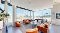Located at 10 Patriot Pkwy in South Weymouth, MA, this thoughtfully designed single-floor community lounge offers a vibrant and inviting atmosphere ideal for gathering and relaxation. The open-concept layout seamlessly connects a fully equipped kitchenette — outfitted with teal cabinetry, stainless steel appliances, and a dining area beneath a statement pendant light — to a comfortable living space anchored by a striking ombre teal accent wall. Hexagonal tile flooring adds a modern, geometric touch throughout.

The living area features mid-century modern furnishings, including leather sofas and accent chairs arranged around a central coffee table, creating a warm and social atmosphere. A dedicated lounge nook showcases a sleek built-in linear fireplace beneath a wall-mounted television, set against a bold gray accent wall for a cozy, refined focal point.

Floor-to-ceiling windows wrap around a bright game area, flooding the space with abundant natural light and offering expansive outdoor views of the surrounding landscape. A full-size shuffleboard table serves as a centerpiece for recreation, complemented by additional seating. The combination of high ceilings, large-format glazing, and curated modern décor gives this community space a sophisticated yet approachable character.
