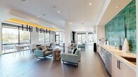 Located at 2525 Paseo Park Rd in Orlando, Florida, this single-floor amenity lounge showcases a thoughtfully designed interior that blends contemporary aesthetics with functional comfort. The open-plan layout is anchored by large floor-to-ceiling windows along multiple walls, flooding the space with natural light and offering views of the surrounding landscaped exterior.

The lounge is divided into several distinct seating zones. A central gathering area features teal upholstered armchairs arranged around organic stone side tables atop large-format dark tile flooring, creating a relaxed yet refined atmosphere. Nearby, a built-in wet bar is accented by a bold teal zellige tile backsplash with warm wood cabinetry, providing a striking visual focal point.

A dining nook showcases a geometric patterned tile floor beneath a dramatic canopy of cylindrical pendant lights suspended from a warm yellow-accented ceiling, accompanied by a long communal table and modern white chairs. Adjacent café-style seating near the windows offers casual high-top tables and upholstered stools ideal for informal gatherings.

A textured sculptural wall with soft, layered paneling adds dimension and warmth to a cozy banquette seating nook with brass wall sconces. Throughout the space, recessed lighting and designer pendant fixtures contribute to a polished, welcoming ambiance.