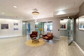This generously sized lobby in Arlington, MA offers an impressive array of amenities spread across a single, thoughtfully designed floor. The entry area sets a welcoming tone with light wood-look tile flooring, a striking pendant chandelier of cascading globe lights, and a curated seating arrangement featuring rich brown leather armchairs, a woven jute rug, and a round coffee table. A curved accent wall adorned with framed historical photographs adds a distinctive architectural element and a nod to local character.

Adjacent spaces flow naturally from the central lobby, including a well-equipped reception and common area with herringbone-patterned flooring, dark wood cabinetry, pendant lighting, and wall-mounted displays. The community lounge area features soft teal-blue walls, barn-style sliding doors, and versatile seating arrangements suited for both social gatherings and quiet relaxation — including a bright, sunlit lounge with a large sectional sofa.

A dedicated game room offers a full-size billiards table and casual seating surrounded by natural light from multiple windows. The fitness center is expansive and fully equipped with cardio machines, strength training equipment, and patterned carpet flooring, all accessed through a glass door entry. Recessed and flush-mount lighting throughout maintains a clean, modern aesthetic across every area of this multifunctional lobby.