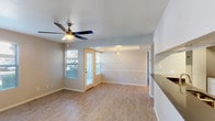This single-floor residence offers a clean, functional layout with a welcoming interior and outdoor access. The main living area is finished with light wood-look flooring that extends throughout, contributing to a bright and cohesive aesthetic. Neutral gray walls provide a versatile backdrop, while a ceiling fan with a built-in light fixture adds both practicality and a subtle decorative touch to the space. A door with sidelights leads directly to the private patio, allowing natural light to filter into the interior and creating a seamless connection between indoor and outdoor areas. A tray ceiling detail in the adjoining dining or transitional area adds a modest architectural element, complemented by a pendant light fixture. Built-in shelving along one wall offers convenient storage or display options. The private patio is framed by textured stucco walls and opens to views of a well-maintained lawn, covered parking carports, and mature trees under clear, sunny skies—suggesting a warm, arid climate setting. The overall ambiance is bright, neutral, and move-in ready.