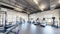 Located at 600 North Avenue in New Rochelle, NY, this fitness center offers a comprehensive workout facility within a spacious, single-floor open layout. The space features an industrial-modern aesthetic, with exposed ceiling ductwork and conduit complemented by clean white walls and durable rubber flooring throughout.

The cardio zone is well-appointed with multiple treadmills and stationary bikes, including a rowing machine, all equipped with digital display screens. The strength training area includes a Rogue power rack loaded with weight plates, adjustable benches, and a cable cross machine, providing a full range of resistance training options. A dedicated free weights section features organized dumbbell racks alongside additional functional fitness accessories such as a medicine ball and resistance equipment.

Natural light fills the space through multiple large windows fitted with roller shades, offering views of the surrounding area. Pendant-style linear LED fixtures supplement the natural light, ensuring the space remains bright and functional at all times. Full-length mirrors along one wall enhance the sense of openness and support proper form during workouts.

Glass doors lead to an adjacent outdoor area, further enhancing the bright, airy atmosphere. The overall design balances a modern industrial character with practical fitness functionality, making this a versatile and well-rounded facility.