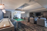 Located at 1490 Ernest W Barrett Pkwy NW in Kennesaw, GA, this single-floor entertainment and game lounge is thoughtfully designed for social interaction and leisure. The space is divided into distinct activity zones, each offering a unique recreational experience. A dedicated home theater area anchors one section, featuring rows of plush, upholstered lounge chairs arranged to face a large wall-mounted flat-screen television flanked by rich, dark wood cabinetry and built-in speakers — ideal for movie nights or group viewing. Adjacent to the theater zone, a billiards area houses a full-size pool table with overhead lighting, complemented by a wall-mounted cue rack for easy access. A sleek shuffleboard table sits against a bold, dark textured accent wall, illuminated by warm pendant lights that add a sophisticated ambiance. A wall-mounted Scrabble board provides a playful, decorative touch throughout. A secondary seating and game table area offers patterned chairs and a dark dining-style table beneath a drum pendant light, suitable for board games or casual gatherings. Bold, patterned carpeting ties the space together, while dark drapery and a drop ceiling with recessed lighting contribute to the lounge's inviting, upscale atmosphere.