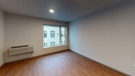 This single-floor unit offers a practical and straightforward layout, combining the kitchen and living area in an open-concept design. The living space is finished with warm hardwood-style flooring and neutral gray walls, creating a clean, versatile backdrop. A large window allows natural light to fill the room while providing views of the surrounding building exterior, and a wall-mounted heating unit ensures year-round comfort.

The kitchen is positioned along one wall and features dark espresso cabinetry with sleek hardware, extending both above and below the countertops for ample storage. A dark granite-style countertop runs the length of the kitchen, paired with a double stainless steel sink and black appliances, including a refrigerator, stove, microwave, and dishwasher. Under-cabinet lighting adds functional illumination to the workspace.

The bathroom maintains a clean, modern aesthetic with crisp white walls and large-format dark gray tile flooring. It includes a walk-in shower with a glass enclosure, a toilet, and a vanity with a dark wood base, all accented by brushed nickel fixtures and a towel bar.