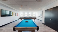 Located at 1352 Randolph Rd in Plainfield, NJ, The Randolph offers a thoughtfully designed single-floor recreational space built for social engagement and entertainment. The game room is spacious and open, featuring light wood-look flooring, a drop tile ceiling with recessed lighting, and crisp white walls that create a bright, clean atmosphere. Multiple windows along the perimeter allow natural light to fill the room, enhancing its welcoming ambiance.

The space is well-equipped with two full-size pool tables dressed in vivid blue felt, a regulation shuffleboard table, and a foosball table — providing a variety of gaming options. A wall-mounted flat-screen television serves as a focal point on one side of the room, flanked by colorful billiard ball-themed artwork that adds a playful, cohesive character to the décor. A dedicated cue rack keeps equipment neatly organized nearby.

A built-in dark shelving unit displays decorative accents and small plants, adding warmth to the otherwise modern aesthetic. Wall-mounted shelves and a chalkboard welcome sign — reading "Welcome to The Randolph!" — contribute a personalized community feel. Seating with teal metal bistro chairs and matching tables is tucked near the windows, offering a casual spot to relax between games. The overall design balances function and style, making this a standout communal amenity.