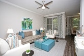 This single-floor residence showcases a thoughtfully designed open-concept layout that seamlessly connects the living and adjoining spaces. The living area centers around rich, dark hardwood flooring that extends throughout, providing a warm and cohesive aesthetic. Crown molding adds a refined architectural detail along the ceiling perimeter, complementing the neutral warm-toned walls.

A ceiling fan with a brushed nickel finish is centrally positioned, providing both functional airflow and a subtle design accent. The living room is generously sized, accommodating a plush sectional sofa paired with a tufted leather ottoman that serves as a central coffee table. A flat-screen television is mounted on the wall opposite, accompanied by a sleek floating media console.

Large windows dressed with layered patterned draperies and horizontal blinds allow abundant natural light to filter into the space, offering views of mature trees outside. A dedicated sitting nook or workspace is visible in the background, featuring a chair and desk arrangement bathed in natural light. An abstract canvas artwork adds a pop of color to the living area, enhancing the overall contemporary yet comfortable ambiance of the home.