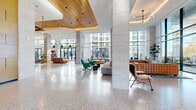Located at 5 Port Imperial Blvd in Weehawken, NJ, this grand single-floor lobby spans an impressive footprint and serves as a dynamic communal hub defined by refined contemporary design. The entrance greets visitors with a dramatic revolving glass door framed by floor-to-ceiling windows that flood the interior with natural light and offer views of the surrounding Port Imperial development. Polished terrazzo flooring extends throughout, providing a sleek, luminous foundation that ties the entire space together.

Soaring ceilings with warm wood slat canopies and recessed lighting create a welcoming, upscale ambiance. Structural columns clad in stacked white brick add texture and rhythm across the open floor plan. A striking reception desk, accented with a translucent blue front panel and a sculptural chandelier above, anchors one section of the lobby. Nearby, a built-in bookshelf wall adds a residential warmth alongside curated lounge seating in varied configurations.

A dedicated game room, enclosed by curved glass block walls, houses a billiards table and large-screen TV against bold emerald-green paneling. Additional social zones feature a foosball table, clustered lounge chairs, a coffee bar with cabinetry, and art installations throughout connecting corridors. The overall atmosphere balances sophisticated architectural detail with an approachable, community-oriented character.