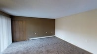 This single-floor space presents a clean and functional interior designed for practical everyday living. The main room is finished with wall-to-wall carpet in a neutral gray tone, providing a comfortable and cohesive foundation throughout. A textured popcorn ceiling adds a classic residential character to the space above.

The room features a distinctive two-tone wall treatment, with a warm brown accent wall complemented by lighter beige surrounding walls, creating a subtle visual contrast. A white baseboard heater runs along the base of the brown accent wall, offering an efficient heating solution integrated seamlessly into the room's perimeter.

A standard interior door is visible along one wall, suggesting access to an adjacent room or closet, contributing to the overall utility of the layout. Multiple electrical outlets are distributed along the walls, supporting flexible furniture arrangements and everyday convenience.

The space is straightforward in its design, emphasizing functionality and ease of use. The neutral color palette and simple finishes make it adaptable to a variety of furnishing styles, while the open floor plan of the main room provides a sense of openness. Overall, the space offers a practical and comfortable environment suited for a range of uses.