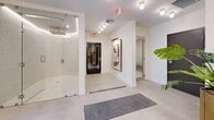 This thoughtfully designed wellness spa offers a calming, nature-inspired environment across a single floor. The entrance opens into a refined lounge area finished with large-format light tile flooring, crisp white walls, and stylish round flush-mount lighting. A woven sunburst mirror, sculptural leather lounge chairs, a tall potted tropical plant, and a full wall of warm wood-toned lockers set an inviting, resort-like tone. A glass-enclosed shower with mosaic tile detailing and an adjacent sauna with a natural wood door provide dedicated amenity facilities. Curated wall art adds a personal touch throughout the common areas. Beyond the lounge, two dedicated treatment rooms offer a tranquil retreat, each bathed in abundant natural light streaming through sheer floor-to-ceiling curtains. The rooms feature warm wood-look plank flooring, massage tables, built-in wood vanities with sinks, and carefully selected botanical artwork. Lush greenery punctuates both rooms, enhancing the organic, wellness-focused atmosphere. The overall design seamlessly blends clean, modern architecture with warm natural materials, creating a cohesive and restorative space ideal for professional wellness and therapeutic services.