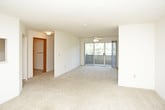 This single-floor residence offers a practical and functional layout across several well-defined spaces. The living area is generously sized, featuring neutral wall-to-wall carpeting and a textured ceiling. A pass-through window connects the living room to the kitchen, providing an open sightline between the two spaces. Natural light enters through a sliding glass door that leads to an exterior balcony or patio area, while a dedicated entry door with a wood finish provides a clear transition from outside. The kitchen is equipped with warm oak cabinetry offering ample storage, white countertops, a stainless steel sink, an electric range, and an over-the-range microwave. Tile flooring grounds the kitchen area. Two bedrooms are each carpeted and feature ceiling fans with light fixtures and windows fitted with horizontal blinds, allowing for natural light and outdoor views of surrounding greenery. The unit includes two bathrooms, both finished with patterned tile flooring. Each bathroom features a tub and shower combination, and one includes a wood-toned vanity cabinet with a single sink and a mirrored medicine cabinet above. Throughout, the space maintains a clean, neutral palette suited for a variety of furnishing styles.