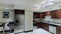 This two-floor space includes a thoughtfully designed kitchen that serves as a standout feature. The kitchen is outfitted with rich, dark espresso-toned wood cabinetry that extends to the upper walls, providing generous storage throughout. Stainless steel appliances, including a side-by-side refrigerator with an in-door water and ice dispenser, a range with an electric cooktop, and an over-the-range microwave, lend a modern, cohesive look. The countertops are finished in granite with subtle variation, complementing the deep tones of the cabinetry. The layout follows an L-shaped configuration, maximizing workspace efficiency. Recessed lighting is installed in the ceiling, supplemented by a track lighting system that offers adjustable illumination across the kitchen area. Light-toned tile flooring runs throughout, contributing to a clean and open feel. The neutral, cream-colored walls keep the space feeling bright and airy. The two-floor layout suggests additional living, sleeping, or functional areas beyond the kitchen, offering a multi-level living experience with room for varied uses across both levels.