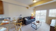 This single-floor residence offers a thoughtfully designed layout across two bedrooms and two full bathrooms. The living room serves as a central gathering space, featuring carpeted flooring, decorative ceiling beams with yellow accent detailing, and a sliding glass door that opens to a private balcony with open outdoor views, allowing natural light to fill the room.

The kitchen is efficiently arranged with warm-toned wood cabinetry extending to the ceiling for generous storage, a stainless steel sink set into a peninsula-style counter, and white appliances including a range, microwave, and refrigerator. The peninsula doubles as a casual dining area.

Both bedrooms are carpeted and receive natural light through large windows with outdoor views. Each room offers comfortable proportions, and at least one bedroom includes a walk-in closet with built-in wire shelving for organized storage.

The two bathrooms each feature wood-tone cabinetry, light countertops, and light hardwood-style flooring. One bathroom includes an extended vanity with wall-mounted cabinetry above, while the other offers a shower-tub combination. Together, the rooms create a cohesive, functional living environment with a neutral, welcoming palette throughout.