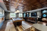Located at 70 Upper Rock Cir in Rockville, MD, this sophisticated game lounge is a well-appointed amenity space designed for social entertainment. The room is immediately distinguished by its ornate coffered copper ceiling, which adds warmth and a dramatic architectural focal point overhead. Rich hardwood flooring runs throughout, complementing the deep navy and charcoal wall tones that give the space a refined, club-like atmosphere.

At the heart of the room sits a full-size billiards table, illuminated by a sculptural pendant light. Adjacent to it, a regulation shuffleboard table stretches along one side of the room, offering additional recreation. A sleek wet bar runs along the back wall, complete with a stone countertop, bar seating with leather stools, and a wall-mounted flat-screen TV framed by textured tile — ideal for entertaining.

A linear fireplace is set into a stone-clad accent wall, flanked by framed artwork, creating an inviting lounge vibe. Striped drapery panels filter natural light from the large windows, while recessed and pendant lighting ensure a well-lit environment at any hour. Glass-partitioned walls connect the space to adjacent amenity areas, enhancing its open, communal character. The overall design balances industrial edge with upscale comfort.