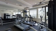 This fitness center offers a clean, well-organized workout environment designed to meet a range of exercise needs. The space is outfitted with a lineup of modern cardio equipment, including treadmills, an elliptical trainer, and a stationary bike, all positioned to take advantage of the natural light streaming in through multiple windows dressed with white horizontal blinds. A dedicated free weights area is visible along one wall, providing options for strength training alongside the cardio equipment. Full-length mirrors line one wall, visually expanding the space and allowing users to monitor their form during workouts. A flat-screen television is mounted on the wall, adding an entertainment option for those using the cardio machines. The ceiling features a combination of recessed lighting and a ceiling fan, ensuring the space remains well-lit and well-ventilated during use. Crown molding details and a coffered ceiling lend a refined architectural touch to the room, elevating the overall aesthetic beyond a standard gym setting. The dark, cushioned flooring provides a practical and durable surface suited for fitness activity. Large windows on multiple walls invite views of surrounding greenery, creating a bright and energizing atmosphere throughout.