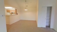 This single-floor space is defined by its vaulted ceilings, which lend an open and airy quality to the main living area. A decorative pendant chandelier hangs centrally, serving as a focal point that enhances the vertical scale of the room. The living area is finished with wall-to-wall carpeting in a warm beige tone, contributing to a comfortable and cohesive aesthetic throughout the space.

A pass-through counter connects the living area to the kitchen, allowing for an open flow between the two spaces. The kitchen features light wood cabinetry and a white appliance finish, visible from the living area, reinforcing the neutral and understated color palette. The counter pass-through also adds a functional element, making the space well-suited for both everyday living and casual entertaining.

A white paneled door is positioned along one wall, providing access to an adjoining room or closet. The walls are finished in a clean, soft white, complementing the natural tones of the flooring and cabinetry. Overall, the space presents a straightforward, functional layout with architectural details that enhance its sense of openness and scale.