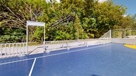 Located at 20950 NW 14th Pl in Miami Gardens, Florida, this outdoor recreational facility offers a well-appointed community gathering and sports space designed to serve active residents of all ages. The centerpiece of the facility is a full multi-sport court surfaced in durable blue and yellow interlocking sport tiles, clearly marked for both basketball and small-sided soccer. The court is equipped with basketball hoops and small soccer goals positioned at each end, making it versatile for a variety of athletic activities. The court is enclosed by sturdy steel cable and rail fencing that provides safety without obstructing sightlines to the surrounding greenery. Bleacher-style seating lines one side of the court, offering spectators a comfortable place to watch. Wide concrete walkways connect the court to the broader grounds, which feature expansive, well-maintained open lawn areas ideal for informal recreation. Picnic tables and benches are situated throughout, providing shaded rest areas and gathering spots. Mature trees border the perimeter, offering natural shade and a lush backdrop against the characteristically bright South Florida sky. The facility enjoys an open, airy ambiance bathed in abundant natural sunlight, creating an inviting environment for community recreation year-round.