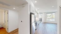 This thoughtfully designed single-floor unit offers a clean, modern aesthetic across seven rooms, with light wood-tone flooring and crisp white walls unifying the entire space. The open-concept kitchen is a standout feature, outfitted with stainless steel appliances including a refrigerator, electric range, and over-the-range microwave, complemented by sleek white cabinetry with bar-pull hardware and white quartz countertops. The kitchen flows seamlessly into the living area, which benefits from large windows and a glass door that invite abundant natural light and offer views of the surrounding neighborhood and hillside scenery. Recessed lighting throughout enhances the bright, airy ambiance. The bedrooms are generously proportioned, featuring the same warm wood-tone flooring, clean white walls, and large windows that frame outdoor views. One bedroom includes a stylish barn-style sliding door, adding a modern design touch. The bathroom is well-appointed with a dark wood-grain vanity, white countertop, a framed mirror, and wood-tone flooring that maintains design continuity throughout the unit. A smart thermostat is also present, reflecting the home's contemporary, tech-forward sensibility. Overall, the space presents a fresh, minimalist environment filled with natural light.