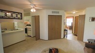 This single-floor residence presents an open and functional layout that connects key living spaces seamlessly. The main living area features light-toned carpeting underfoot and a ceiling fan with integrated lighting, providing both comfort and air circulation. A prominent bi-fold closet offers generous built-in storage, a practical and central feature of the space.

Adjacent to the living area, the kitchen is visible and accessible, finished with white cabinetry, white appliances including a range and dishwasher, and light-colored countertops. The kitchen benefits from under-cabinet lighting and overhead cabinetry that maximizes storage capacity. A partial wall separates the kitchen from the living space, maintaining an open feel while defining each area.

A hallway leads to additional rooms, with a glimpse of a sunlit space at the end suggesting a room with good natural light from an exterior window. The overall color palette throughout is neutral and light, with white and warm beige tones dominating the walls, ceilings, and floors, contributing to a bright and airy ambiance. The single-floor layout ensures easy accessibility and a cohesive flow between all areas of the residence.