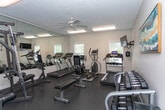 Located at 117 Teakwood Dr in Greensboro, North Carolina, this single-floor fitness center is a well-equipped exercise facility designed to support a comprehensive workout routine. The space features durable rubber flooring throughout, providing a safe and functional surface for all types of exercise.

The cardio section is outfitted with multiple treadmills, an elliptical trainer, and a stationary cycle, offering a variety of options for cardiovascular workouts. A wall-mounted flat-screen television is positioned for easy viewing during cardio sessions. For strength training, the facility includes a multi-station cable machine with an adjustable bench, a dedicated free weights area with a tiered dumbbell rack, and additional benches for various exercises.

Full-length mirrors line one wall, enhancing the sense of space while allowing users to monitor their form during workouts. Bright flush-mounted fluorescent lighting ensures the room is well-lit at all times, complemented by natural light streaming in through several windows fitted with blinds. Colorful wall art adds a touch of vibrancy to the otherwise clean, neutral-toned interior. A dedicated entry door provides easy access to the space. Overall, the fitness center presents a clean, organized, and functional environment suited for both cardio and strength training needs.