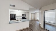 This well-designed single-floor residence offers a bright, open layout with a seamless flow between the main living areas. The kitchen is outfitted with white and light wood cabinetry, a gray countertop peninsula with a double-basin sink, stainless steel appliances including a microwave and refrigerator, and a black electric range. The peninsula opens directly to the living space, making it ideal for everyday living and entertaining. The living room is a standout space, featuring vaulted ceilings, recessed lighting, a classic white-surround fireplace, and large windows fitted with horizontal blinds that invite generous natural light. A sliding glass door provides direct access to an outdoor area. Hardwood-style flooring runs throughout the main living spaces, adding warmth and continuity. The bedroom features plush carpeting and a textured ceiling, with a door leading to a connected bathroom. The bathroom is equipped with a wide vanity, gray composite countertop, light wood cabinetry with brushed nickel hardware, an oversized mirror with Hollywood-style lighting, and wood-style flooring. A hallway connects the rooms, with closet storage accessible via classic bi-fold doors. The overall ambiance is clean, neutral, and move-in ready.