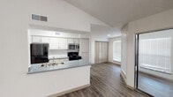 This well-designed single-floor residence offers a bright, open layout with a seamless flow between the main living areas. The kitchen is outfitted with white and light wood cabinetry, a gray countertop peninsula with a double-basin sink, stainless steel appliances including a microwave and refrigerator, and a black electric range. The peninsula opens directly to the living space, making it ideal for everyday living and entertaining. The living room is a standout space, featuring vaulted ceilings, recessed lighting, a classic white-surround fireplace, and large windows fitted with horizontal blinds that invite generous natural light. A sliding glass door provides direct access to an outdoor area. Hardwood-style flooring runs throughout the main living spaces, adding warmth and continuity. The bedroom features plush carpeting and a textured ceiling, with a door leading to a connected bathroom. The bathroom is equipped with a wide vanity, gray composite countertop, light wood cabinetry with brushed nickel hardware, an oversized mirror with Hollywood-style lighting, and wood-style flooring. A hallway connects the rooms, with closet storage accessible via classic bi-fold doors. The overall ambiance is clean, neutral, and move-in ready.