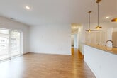 This single-floor residence offers a clean, contemporary design throughout. The open-concept living and dining area is finished with warm wood-style flooring, crisp white walls, and recessed lighting. A set of sliding glass doors fills the space with natural light and provides outdoor views, while a window with blinds adds additional brightness. The kitchen is positioned adjacent to the living area and features white shaker cabinetry, quartz-style countertops, a mosaic tile backsplash, and a full suite of stainless steel appliances including a refrigerator, electric range, and microwave. A raised breakfast bar separates the kitchen from the main living space, complemented by pendant lighting overhead. A large louvered bi-fold closet provides ample storage near the kitchen entry. The bedrooms are carpeted with neutral-toned, textured carpet tiles, offering a quieter, softer feel. Each bedroom benefits from windows that allow natural light and outdoor views. The bathroom includes a dark wood vanity with a light countertop, tile flooring, and a full soaking tub with a shower, maintaining the unit's clean and functional aesthetic throughout.