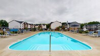 Located at 5695 Cherry Bottom Rd in Columbus, Ohio, this outdoor amenity space is centered around a generously sized resort-style swimming pool that serves as the focal point of the community. The pool features a classic rectangular design with a bright turquoise finish, complemented by a wide concrete deck that provides ample space for lounging and socializing. Surrounding the pool are numerous lounge chairs and patio furniture sets shaded by umbrellas, offering comfortable spots for relaxation. The deck is enclosed by a black iron perimeter fence, providing both safety and a clean visual boundary. In the background, multi-story residential buildings with traditional architectural detailing, pitched dark roofs, and white and gray exteriors frame the space, giving it a vibrant community atmosphere. Mature trees and greenery line the perimeter, adding a natural backdrop to the setting. The overall ambiance is that of a well-appointed, community-focused outdoor retreat designed for warm-weather enjoyment and social gathering.