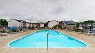 Located at 5695 Cherry Bottom Rd in Columbus, Ohio, this outdoor amenity space is centered around a generously sized resort-style swimming pool that serves as the focal point of the community. The pool features a classic rectangular design with a bright turquoise finish, complemented by a wide concrete deck that provides ample space for lounging and socializing. Surrounding the pool are numerous lounge chairs and patio furniture sets shaded by umbrellas, offering comfortable spots for relaxation. The deck is enclosed by a black iron perimeter fence, providing both safety and a clean visual boundary. In the background, multi-story residential buildings with traditional architectural detailing, pitched dark roofs, and white and gray exteriors frame the space, giving it a vibrant community atmosphere. Mature trees and greenery line the perimeter, adding a natural backdrop to the setting. The overall ambiance is that of a well-appointed, community-focused outdoor retreat designed for warm-weather enjoyment and social gathering.