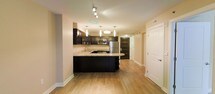 This well-appointed single-floor residence showcases a thoughtfully designed open-concept layout that maximizes both functionality and aesthetic appeal. The kitchen serves as the centerpiece of the space, featuring bold dark cabinetry that creates a striking contrast against the light granite countertops. A generously sized kitchen island anchors the room and provides additional workspace and seating, making it ideal for both everyday meals and entertaining. Stainless steel appliances, including a refrigerator, microwave, and range, add a polished, contemporary finish. Overhead, a combination of stylish pendant lights and a track lighting system illuminate the kitchen with a warm, inviting glow. The warm-toned hardwood flooring flows seamlessly throughout, lending continuity and a sense of spaciousness to the entire floor. Neutral-painted walls complement the overall color palette, keeping the atmosphere bright and welcoming. A hallway leading from the kitchen connects to additional areas of the residence, with clean white doors and subtle architectural details reinforcing the modern design sensibility. The cohesive blend of dark, light, and warm tones throughout creates a balanced and sophisticated living environment.