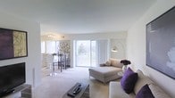 This single-floor residence offers a clean, modern layout designed to maximize comfort and natural light. The living area is generously proportioned, featuring plush wall-to-wall carpeting, crisp white walls, and smooth ceilings that lend a sense of openness throughout the space. A large sliding glass door serves as a focal point of the living room, flooding the interior with natural light and providing direct access to a private balcony that overlooks a lush, green outdoor landscape — a standout feature of the home.

The living room is thoughtfully arranged, with ample space for comfortable seating and a stylish arc floor lamp that adds a contemporary touch. Accent pillows in deep purple bring a pop of color against the neutral tones of the furnishings and walls.

Adjacent to the living area, the dining space is visible through a partial wall opening, creating a connected yet distinct area. The dining space benefits from its own window, ensuring it remains bright and welcoming. The kitchen is accessible nearby, completing the practical flow of the common areas.

Overall, the space presents a bright, neutral palette with subtle modern accents, offering a serene and functional living environment well-suited for everyday comfort.