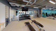This contemporary fitness center offers a comprehensive workout environment designed to accommodate a wide range of exercise needs. The space features an industrial-style aesthetic, with an exposed concrete ceiling accented by visible ductwork and pipe systems, all illuminated by sleek linear lighting fixtures throughout.

The main floor is divided into distinct workout zones. The cardio area is lined with modern elliptical machines, stationary bikes, and treadmills, positioned along expansive floor-to-ceiling glass doors that open to an outdoor patio area. Natural light floods this zone, offering views of an exterior climbing wall and outdoor seating. A dedicated free weights section includes a full dumbbell rack with adjustable benches, complemented by wall-mounted mirrors and mounted flat-screen TVs for entertainment.

A separate strength training area, distinguished by a vibrant teal accent wall and mirrored surfaces, houses a selection of selectorized resistance machines suited for targeted muscle training. The flooring throughout is a durable speckled rubber material, practical and resilient for high-traffic gym use.

Additional amenities visible within the space include storage shelving, a whiteboard, and interior glazed doors connecting separate functional zones. The overall layout is open, spacious, and thoughtfully organized to support diverse fitness routines in a clean, modern setting.