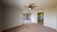 This single-floor residence offers a well-organized layout across several interconnected rooms, as seen in the 3D floor plan. The living area is generously proportioned, featuring neutral-toned carpet, white walls, and a ceiling fan with a light fixture that provides both airflow and ambient lighting. A ceiling vent ensures consistent climate control throughout. An open passage connects the living space to the kitchen, which is equipped with dark cabinetry featuring gold hardware, laminate countertops, a gas range, dishwasher, and a refrigerator — all set atop light vinyl flooring.

The bedrooms are carpeted and offer ample space, each with dedicated closet storage. One bedroom features a spacious walk-in closet with dual hanging rods on either side, while another includes a reach-in closet with built-in shelving. A large window in one bedroom frames a scenic view of treetops, bringing in natural light. The bathroom includes a tub-shower combination with tiled surround, a wood-vanity with a white countertop sink, a large mirror, and blue mosaic tile flooring. A separate water closet provides added privacy.