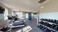 This fitness center is a thoughtfully designed, single-floor facility built to support a wide range of workout routines. The interior is finished with a distinctive two-tone wall treatment — warm gray on the upper half and a crisp light teal band running at mid-height — creating a clean, energizing atmosphere throughout the space. Durable modular carpet tiles in a charcoal multi-tone pattern cover the floors, providing both traction and comfort underfoot.

The main workout area is generously sized and lined with multiple windows on three sides, flooding the room with natural light and offering views of the surrounding community grounds. Ceiling fans are positioned throughout to maintain airflow during workouts. Recessed lighting supplements the natural light for a consistently bright environment.

Cardio equipment is prominently featured, including a row of treadmills and elliptical trainers from Spirit Fitness, positioned to face the windows. A wall-mounted flat-screen TV is available for entertainment during workouts. Strength training options include a multi-functional cable machine, a full dumbbell rack with an extensive weight range, a TRX suspension training system, a spin bike, a stability ball, a workout bench, and a plyometric jump box. An exterior door provides direct access to an outdoor pool area, further enhancing the facility's amenities.