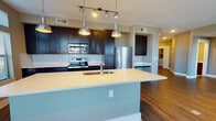 This well-appointed single-floor residence offers a thoughtfully designed open-concept layout that seamlessly connects the kitchen, dining, and living areas. The kitchen is a standout feature, showcasing rich dark cabinetry, a classic white subway tile backsplash, stainless steel appliances including a range, microwave, and refrigerator, and a generous island with a light quartz countertop and undermount sink. Sleek industrial-style pendant lights hang above the island, complementing the track lighting overhead. Warm hardwood-style flooring flows throughout the main living areas, adding cohesion and elegance to the space. The living area is bathed in natural light through expansive floor-to-ceiling windows and glass doors that open to a balcony, offering outdoor views and an airy atmosphere. Both bedrooms feature large multi-pane windows, dark hardwood-style flooring, ceiling fans, and neutral wall tones that create a calm, restful environment. The two full bathrooms are finished with tile flooring, dark vanity cabinetry, light quartz countertops, and full soaking tub and shower combinations. The overall design balances contemporary style with functional living, presenting a clean and modern aesthetic throughout.