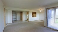 This single-floor residence offers an open and functional layout designed to maximize usable space. The main living area is finished with neutral wall-to-wall carpeting, contributing to a clean and understated aesthetic. A ceiling fan with integrated lighting is centrally positioned, providing both air circulation and ambient illumination throughout the space.

A defining architectural feature is the pass-through window connecting the living area to the kitchen, allowing natural sightlines between the two spaces while maintaining a degree of separation. The kitchen is equipped with wooden cabinetry in a warm tone, along with standard appliances including a refrigerator and range. The kitchen flooring transitions to tile, distinguishing it from the carpeted living area.

Adjacent to the main living space, built-in open shelving units offer practical storage and display options. A doorway nearby provides access to an additional room, adding to the overall versatility of the floor plan. The transition zone between rooms features tile flooring, reinforcing a clear delineation between functional areas.

Overall, the space maintains a bright, neutral palette with white walls and light-toned finishes, creating a straightforward and adaptable environment.