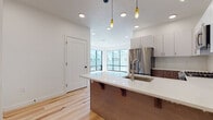 This single-floor unit showcases a contemporary design with clean lines and a cohesive neutral palette throughout. The open-concept kitchen and living area serves as the heart of the space, anchored by a substantial island with a white quartz countertop, an undermount sink, and seating overhang supported by wood brackets. The kitchen features a two-tone cabinetry scheme, pairing white upper cabinets with warm wood-grain lower cabinets, complemented by a white subway tile backsplash and a full suite of stainless steel appliances, including a gas range, over-the-range microwave, and refrigerator. Pendant lighting with Edison-style bulbs adds a stylish accent above the island. Light hardwood flooring extends seamlessly through the kitchen and living areas, while the living room benefits from expansive floor-to-ceiling windows and a sliding glass door that bring in abundant natural light and outdoor views. A modern sliding door connects the living area to the kitchen. The bedrooms feature plush carpeting and recessed lighting, with one room offering a window framing outdoor views. The bathroom is finished with a wood-grain vanity, white quartz countertop, a large mirror, and a full soaking tub with shower combination.