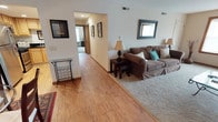 This single-floor residence offers a thoughtfully designed layout with a variety of functional living spaces. The living room is a comfortable and inviting area, featuring plush carpeting, a large picture window that draws in natural light, and ample space for both relaxing and entertaining. A substantial shelving and entertainment unit anchors one wall, providing generous storage and display space. The kitchen is well-equipped with light wood cabinetry, dark granite countertops, stainless steel appliances including a gas range and dishwasher, and recessed ceiling lighting, all set over hardwood-style flooring. Adjacent to the kitchen, the dining area accommodates a round glass-top table beneath a ceiling fan with light fixture. A hallway with hardwood-style flooring connects the main living areas to the private rooms. The primary bedroom is generously sized with soft carpet, a ceiling fan, and a window for natural light. A second bedroom doubles as a versatile home office, also featuring carpet, a ceiling fan, and a wide window. The full bathroom is finished with a dark granite vanity, tile flooring, a tub/shower combination, and a textured ceiling. Throughout the home, neutral tones, consistent trim work, and ceiling fans contribute to a cohesive and comfortable living environment.