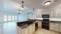 This single-floor residence showcases a thoughtfully designed layout with high-quality finishes across every room. The kitchen serves as a central highlight, featuring glazed antique-white cabinetry with detailed molding, dramatic dark granite countertops with gold veining, a large undermount double sink, and stainless steel appliances including a gas range with double oven and built-in microwave. Light wood-look tile flooring runs throughout the main living areas, complemented by decorative iron pendant lighting and chandelier fixtures. A built-in hutch with glass-front cabinetry and open shelving anchors the dining area, which opens to a spacious living room with access to an outdoor balcony, allowing natural light to pour in. The bedrooms are carpeted and generously sized, each featuring wide windows with outdoor views and ceiling fans. Two well-appointed bathrooms feature large-format marble-look tile, dark granite vanity tops, white shaker cabinetry with oil-rubbed bronze hardware, and glass-enclosed showers. A dedicated laundry room includes white cabinetry, a utility sink with dark granite countertop, and an in-unit washer.