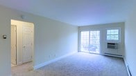 This single-floor space features a spacious living area finished with wall-to-wall plush carpeting in a neutral tone, contributing to a clean and comfortable atmosphere throughout. The walls are painted in a soft, light hue that enhances the sense of openness and pairs well with the bright white baseboards and trim that frame the room. A large sliding glass door serves as a prominent architectural feature, allowing abundant natural light to flood the interior while providing direct access to an outdoor space, where greenery is visible beyond the glass. The arched doorway on one side adds a subtle architectural detail, offering a soft transition to adjoining areas of the space. The overall layout is open and uncluttered, lending flexibility for a variety of furniture arrangements. The combination of natural light, neutral finishes, and carpeted flooring creates a calm, inviting ambiance suited for everyday living.