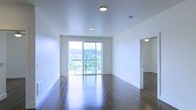 This single-floor space is defined by its clean, modern design and an abundance of natural light. The main living area features rich, dark hardwood flooring that contrasts sharply with the crisp white walls and smooth white ceiling, creating a sleek, contemporary aesthetic. Recessed flush-mount ceiling lights provide even ambient illumination throughout the space.

The most prominent feature of the living area is the large floor-to-ceiling sliding glass door, which opens onto a balcony and frames an expansive view of a green, urban landscape below. This glazed wall floods the interior with natural light and creates a strong visual connection between the indoors and the outdoors, contributing to an airy, open ambiance.

The layout is open and unobstructed, offering flexible arrangement possibilities. A doorway visible to the right leads to an additional room or hallway, suggesting a functional separation of spaces within the unit. The overall design prioritizes simplicity, with minimal architectural detailing allowing the natural light and outdoor views to serve as the primary focal points of the space.