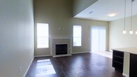 This two-floor space offers an impressive and well-appointed layout designed for comfortable living. The main living area immediately captures attention with its soaring vaulted ceilings that create a dramatic sense of openness and volume. Rich, dark hardwood flooring runs throughout, adding warmth and a polished aesthetic to the space.

A standout feature of the living area is the classic fireplace, framed by a clean tile surround that extends to a modest hearth — serving as a natural focal point for the room. Neutral, earthy wall tones complement the flooring and contribute to a calm, inviting atmosphere throughout.

Natural light is abundant, with multiple windows positioned along the walls, including a large sliding glass door fitted with vertical blinds that provides access to an outdoor area. The generous window placement ensures the space remains bright and airy throughout the day.

An open upper-level overlook is visible from the main living area, reinforcing the two-story design and adding architectural depth. A ceiling vent is also visible, indicating a central HVAC system for year-round climate control.

Overall, this space combines functional design with elevated architectural details, making it a bright, spacious, and comfortable environment suited for a variety of living arrangements.