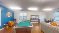 This single-floor community space is thoughtfully designed to foster social interaction and recreational activity across several distinct functional zones. The open layout is anchored by warm hardwood-style flooring throughout, with neutral white walls and flush panel ceiling lighting that keep the atmosphere bright and welcoming.

The main lounge and game area is generously proportioned, centered around a full-size pool table set atop the hardwood floor. Comfortable seating, including a plush gray sofa and vibrant orange accent chairs, creates an inviting gathering spot alongside a wall-mounted flat-screen TV. A decorative console table with a mirror adds a refined touch to one wall, while multiple windows fitted with blinds allow natural light to filter into the space.

A dedicated dining area features a round table with black cross-back chairs, positioned beside a large window overlooking greenery outside. Adjacent to this is a well-equipped kitchenette with dark espresso cabinetry, granite-style countertops, a sink, a microwave, and a coffee bar station stocked for everyday use.

A standout feature is the built-in craft and activity station, complete with an expansive wooden worktable and a large pegboard wall for organizing supplies and tools. Together, these areas make this space highly functional for both leisure and creative pursuits.