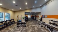 This well-equipped fitness center is organized across a single floor, offering distinct zones for cardio, strength training, and free weights. The layout flows from a free weights area near the entrance through an open passage into a larger cardio and machine room beyond, providing a sense of depth and generous floor space throughout.

The entrance area is visually striking, featuring a bold arched doorway framed in deep orange against a teal accent wall, with a metallic-paneled lower wall section adding an industrial touch. Wall-mounted pull-up brackets are also positioned in this zone alongside a dumbbell rack, adjustable bench, and a cable machine.

Moving into the main area, a rack of medicine balls and a weight bench are positioned alongside cardio equipment. The rear room houses a row of treadmills, ellipticals, and cable strength machines lined along the walls, with natural light provided by a bank of windows. Ceiling fans are distributed throughout for air circulation, complemented by recessed lighting across both zones.

A consistent patterned carpet in neutral tones covers the entire floor, while a bold orange horizontal stripe runs along the walls, unifying the space visually. Posted fitness guidelines and a hand sanitizer station reflect attention to facility management.